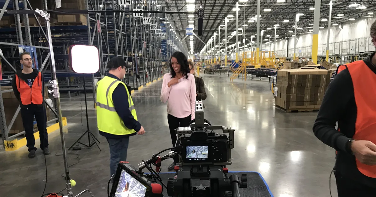 A presenter on camera in a fulfillment warehouse, with the camera rig and monitor in the foreground and crew in safety vests