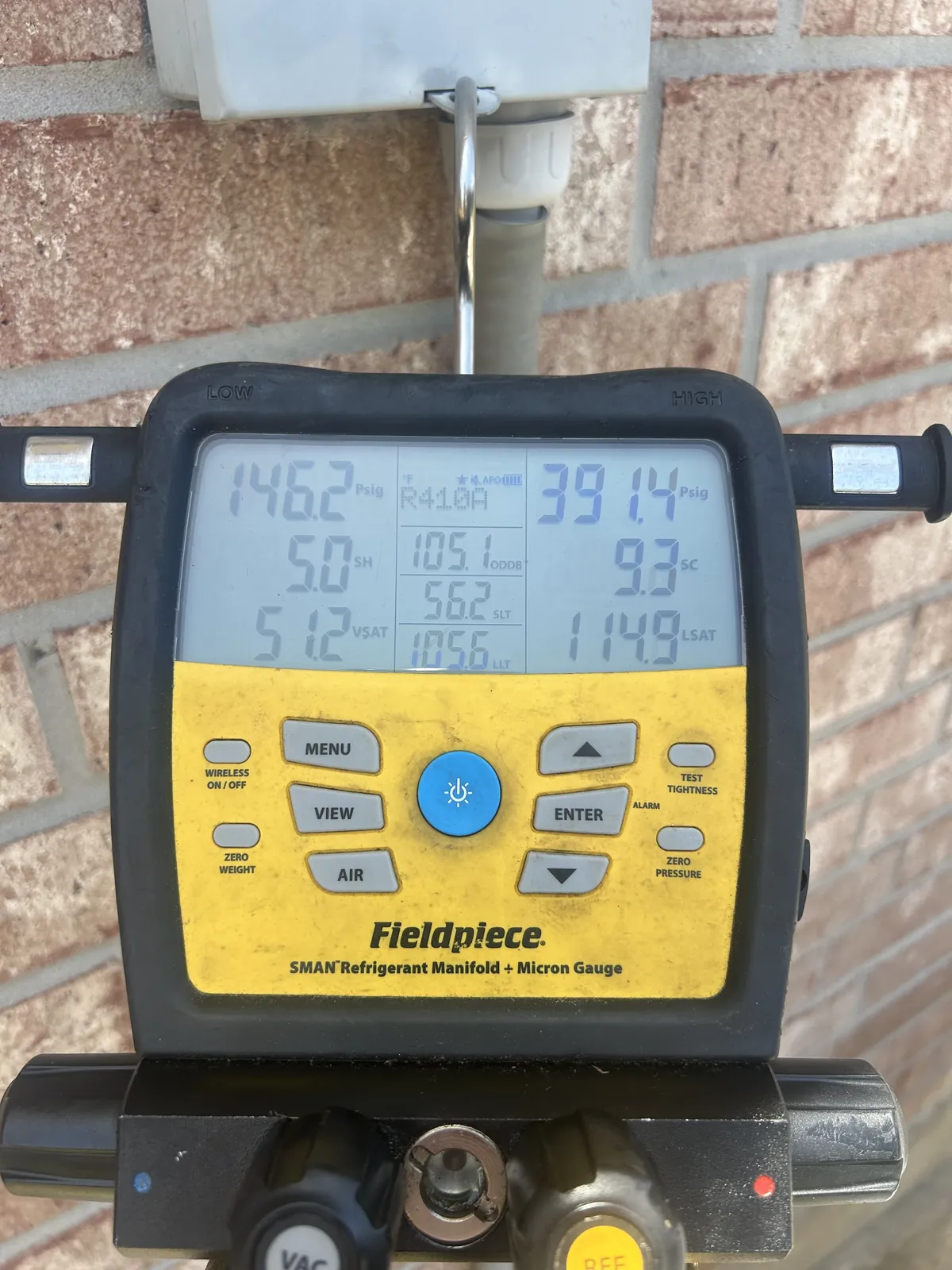 A digital refrigerant manifold gauge hanging on a brick wall, showing live low-side and high-side pressure readings.
