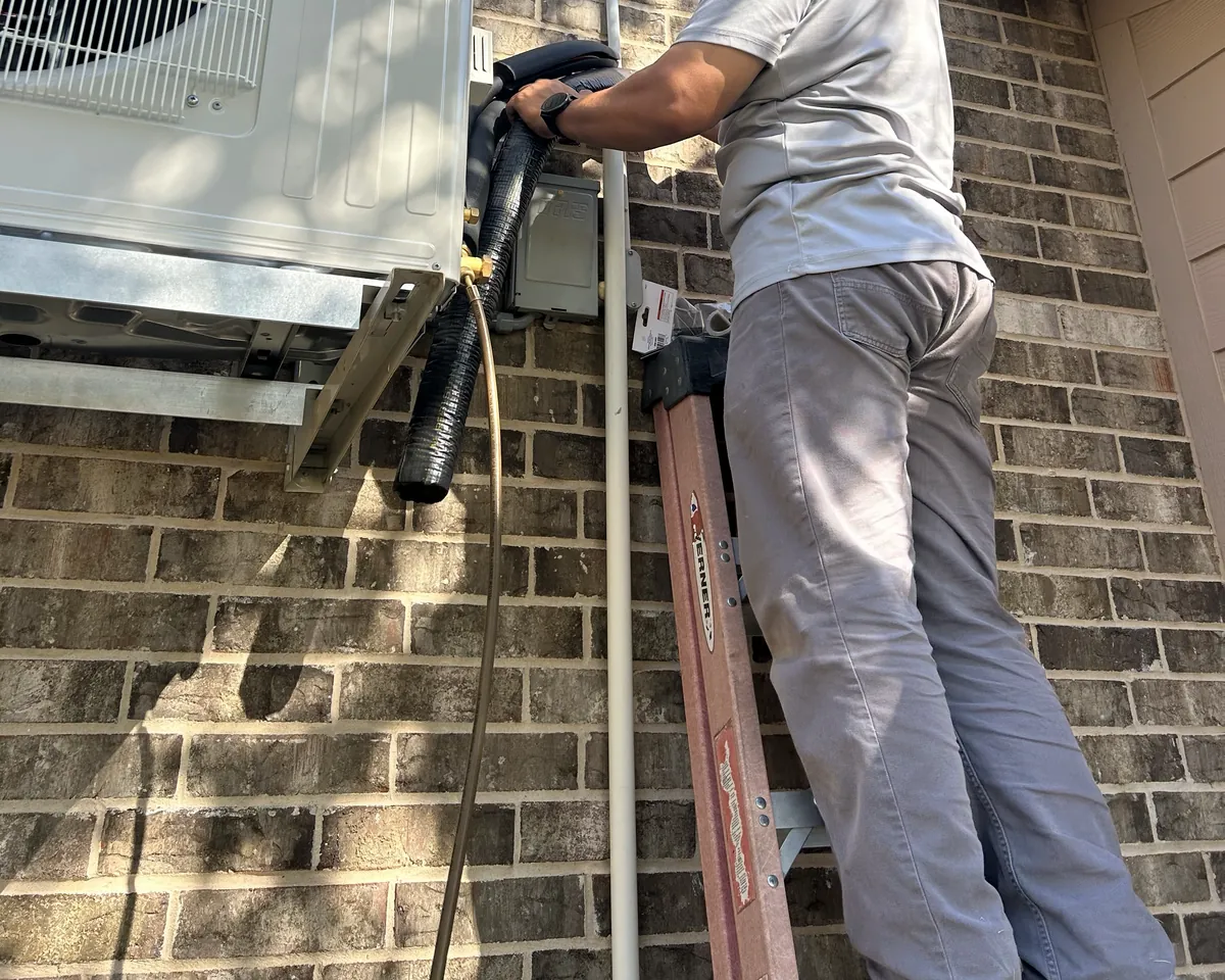 A technician on a ladder connecting the line set to a wall-mounted mini-split outdoor unit on a brick house.