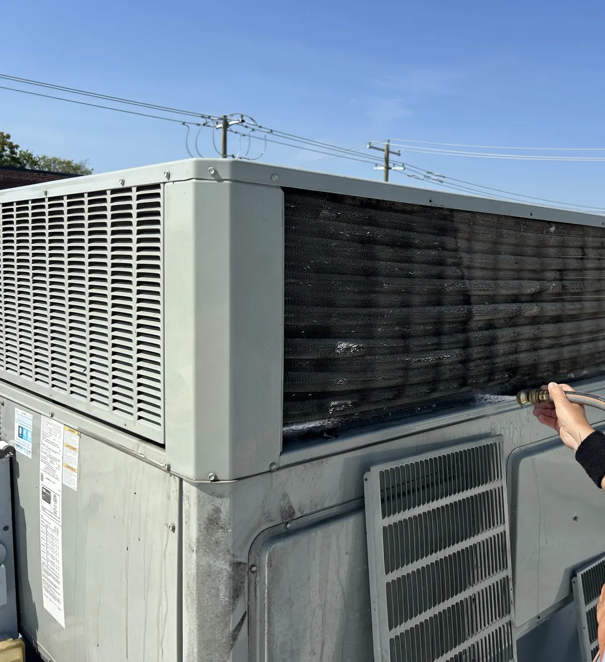 A technician's hand rinsing the condenser coil of a rooftop package unit with a hose.