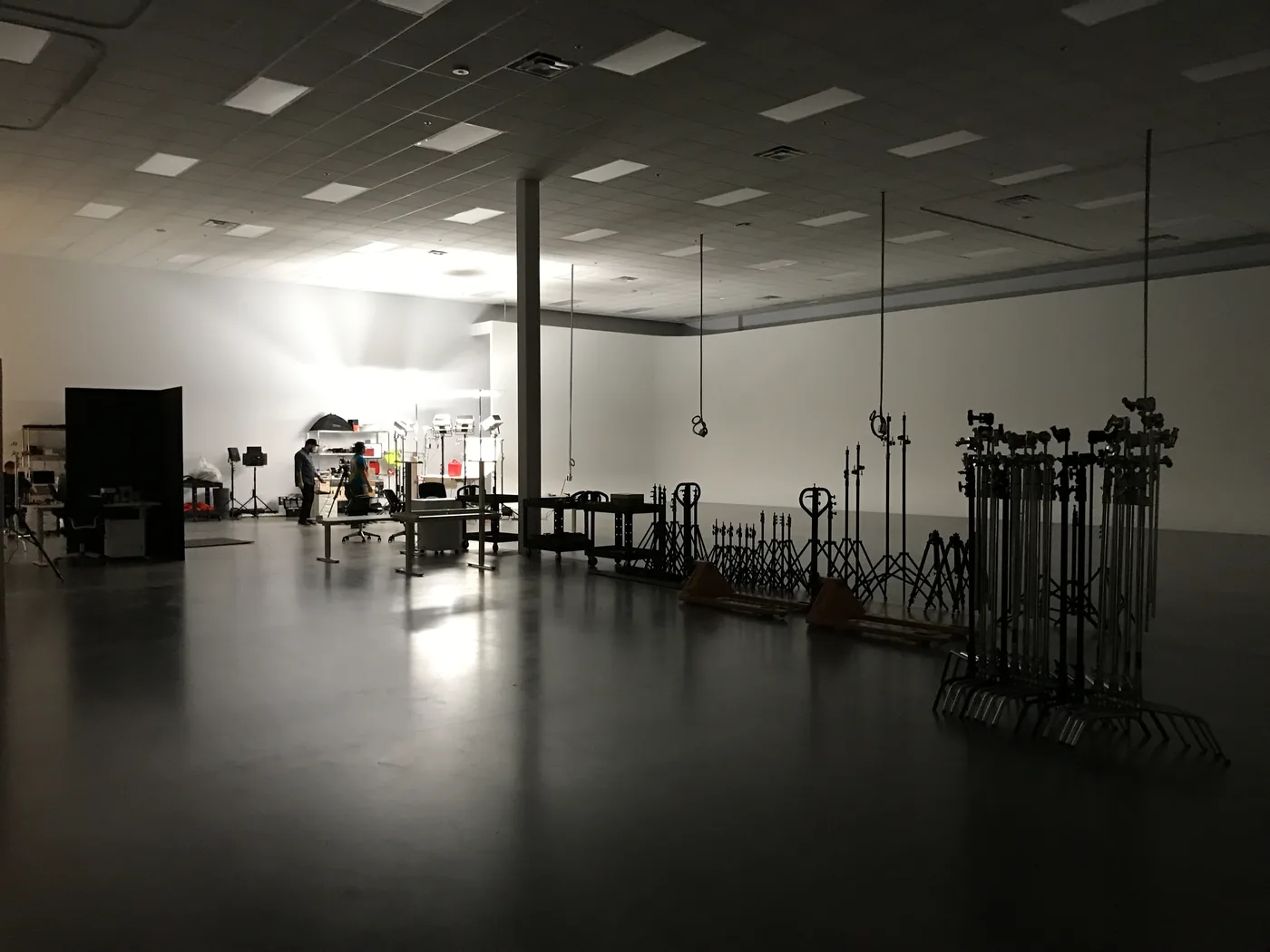 A large, dimly lit production studio with rows of light stands on the floor and two crew members working at the far end