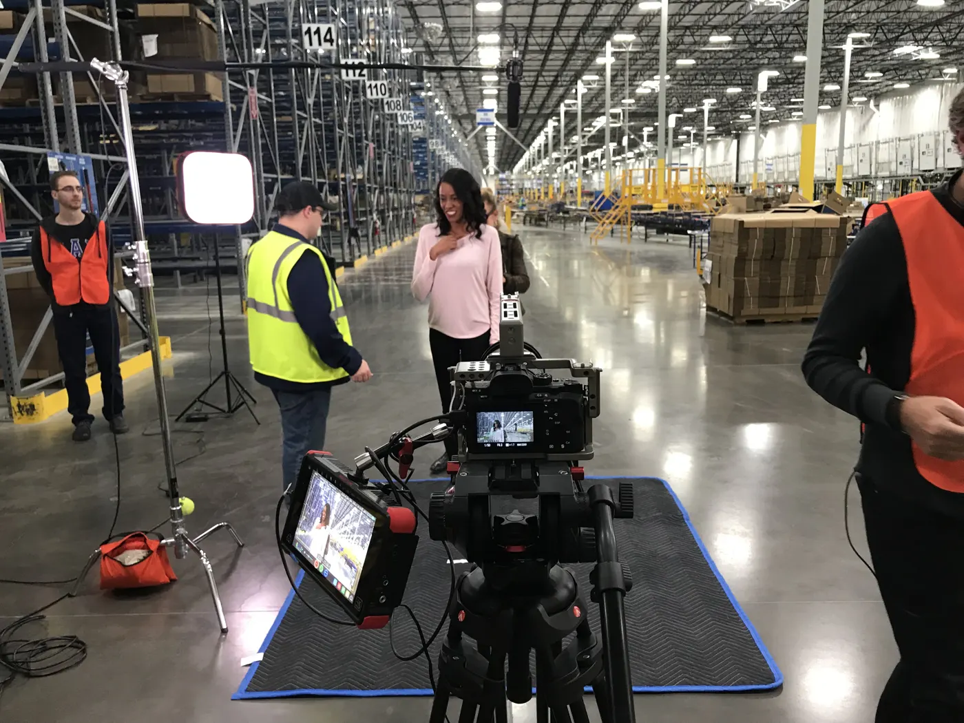 A presenter on camera in a fulfillment warehouse, with the camera rig and monitor in the foreground and crew in safety vests