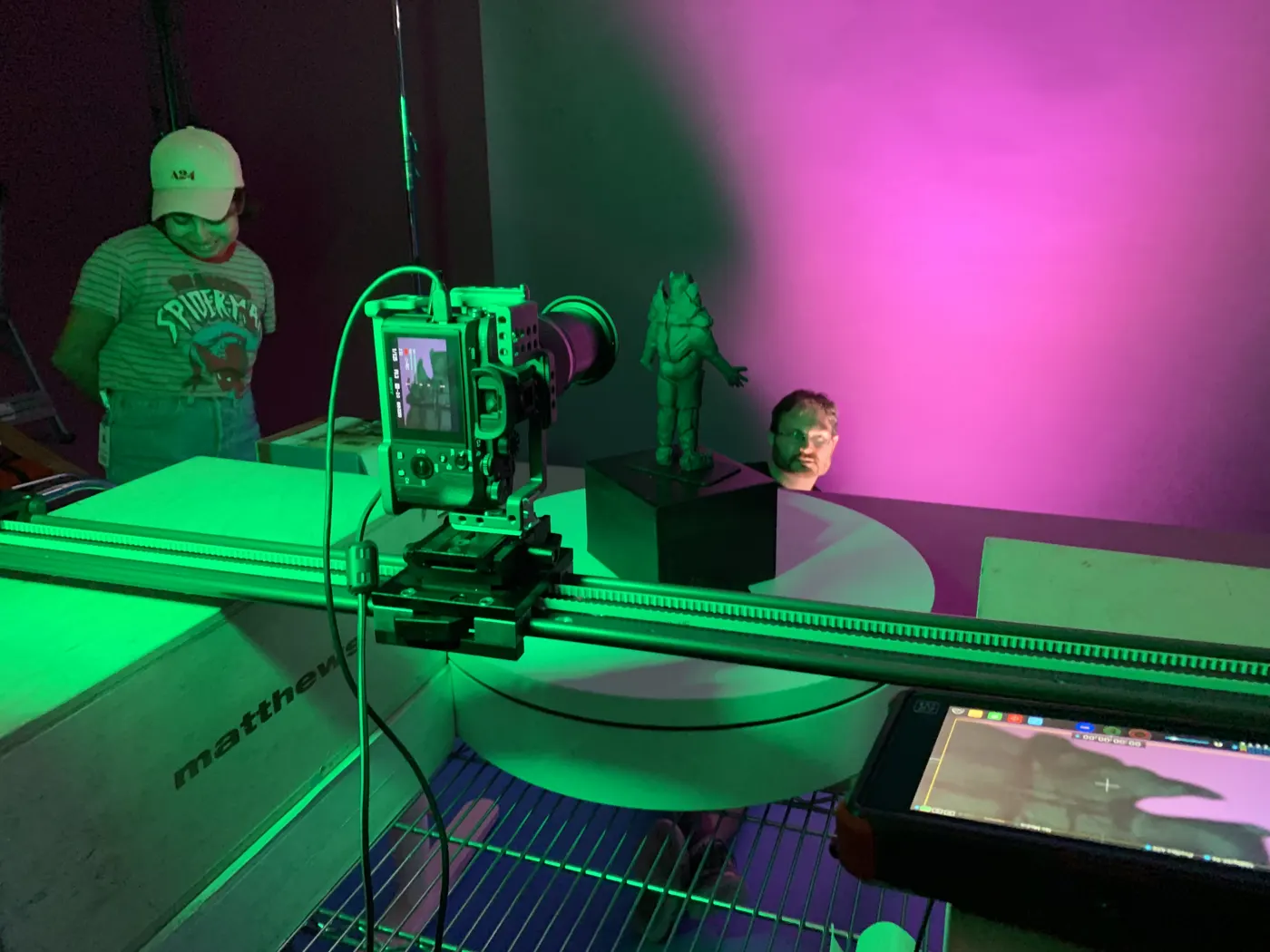 Camera on a slider filming a figurine under green and magenta light, with two crew members watching